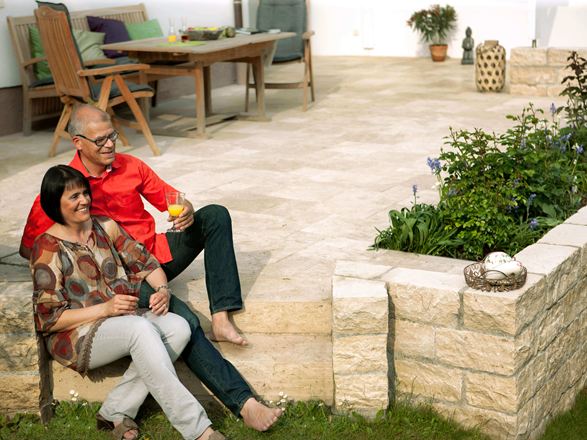 Couple assis sur une terrasse avec des dalles en travertin Medium SELECT et un mur en pierre naturelle, avec en arrière-plan des meubles de jardin en bois.