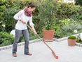 Une femme nettoie une terrasse en granit avec un balai, en arrière-plan un jardin naturel