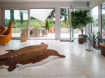 Baie vitrée séjour tapis en peau de bête plante fauteil orange carrelage Vanilla Rustica travertin