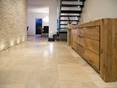 Le carrelage Medium Line, pour une pièce spacieuse. Un sol en travertin sur la droite un commode en bois en arrière plan un mur sur la gauche et des escaliers noirs à droite au fond la cuisine