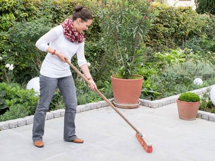 Une femme nettoie une terrasse en granit avec un balai, en arrière-plan un jardin naturel